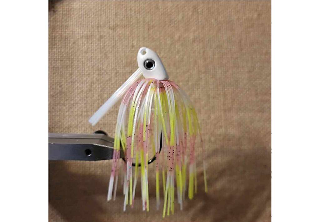 A multicolored fishing lure with a white head and a skirt made of strands of yellow, pink, and white, attached to a metal Fly Tying vice.